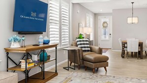 A modern living room with a flat-screen TV, a wooden console table, a leather armchair, a dining area with a table and chairs, and a large window with white shutters. - Coastal Charm Estate | Pool, Golf + Luxury Style! (Selbyville)
