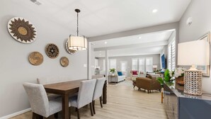 A modern kitchen with a dining area, featuring a wooden table and chairs, a hanging light fixture, and decorative wall art. - Coastal Charm Estate | Pool, Golf + Luxury Style! (Selbyville)