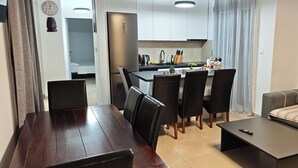 A modern living room with a dark wooden dining table, black chairs, a grey sofa, and a small coffee table. A kitchen with white cabinets and a stainless steel refrigerator is visible in the background.