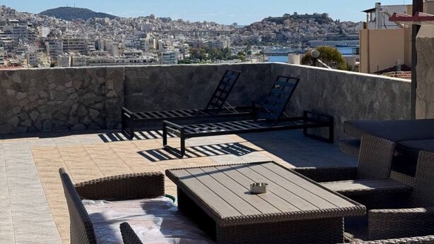 A rooftop terrace with wicker furniture, a stone wall, and a view of a cityscape and hills.