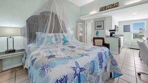 Une chambre à coucher avec un lit sous une moustiquaire, un bureau avec une chaise, un coin cuisine et une salle de bain visible à travers une porte ouverte.