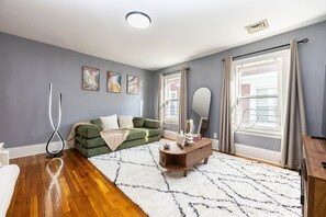 A modern living room with a green sofa, a white rug, a wooden side table, and a large window with curtains. - Cozy 3BD/1BA in Boston | Franklin Zoo, Boston Comm (Boston)