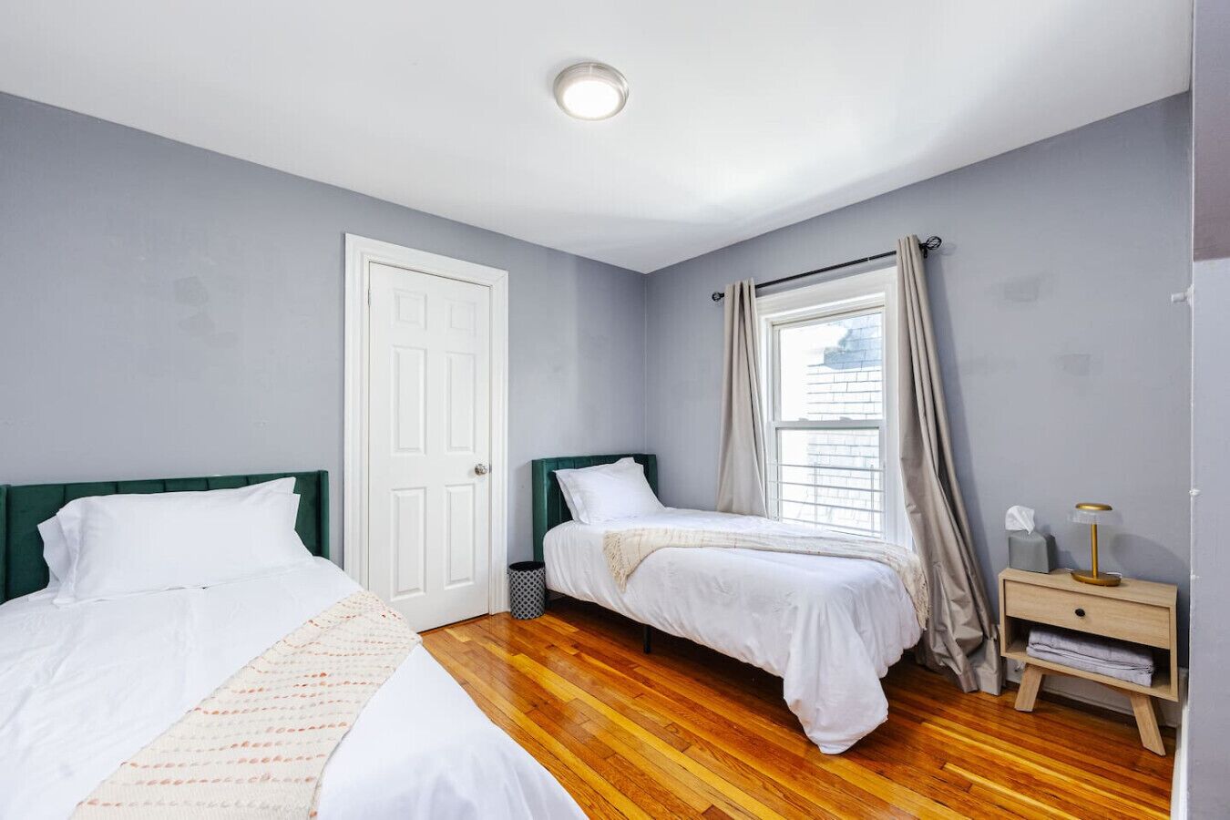 Une chambre avec deux lits, un plancher en bois, une porte blanche et une fenêtre avec des rideaux.