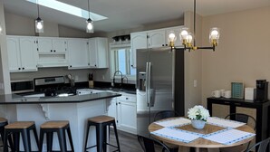 A modern kitchen with white cabinets, a central island with stools, a dining table with chairs, and a chandelier. - Beach House w/ Huge Deck Amazing Views!! Kayaks, Swim, Fish! Golf Cart Rental! (Salem Lakes)