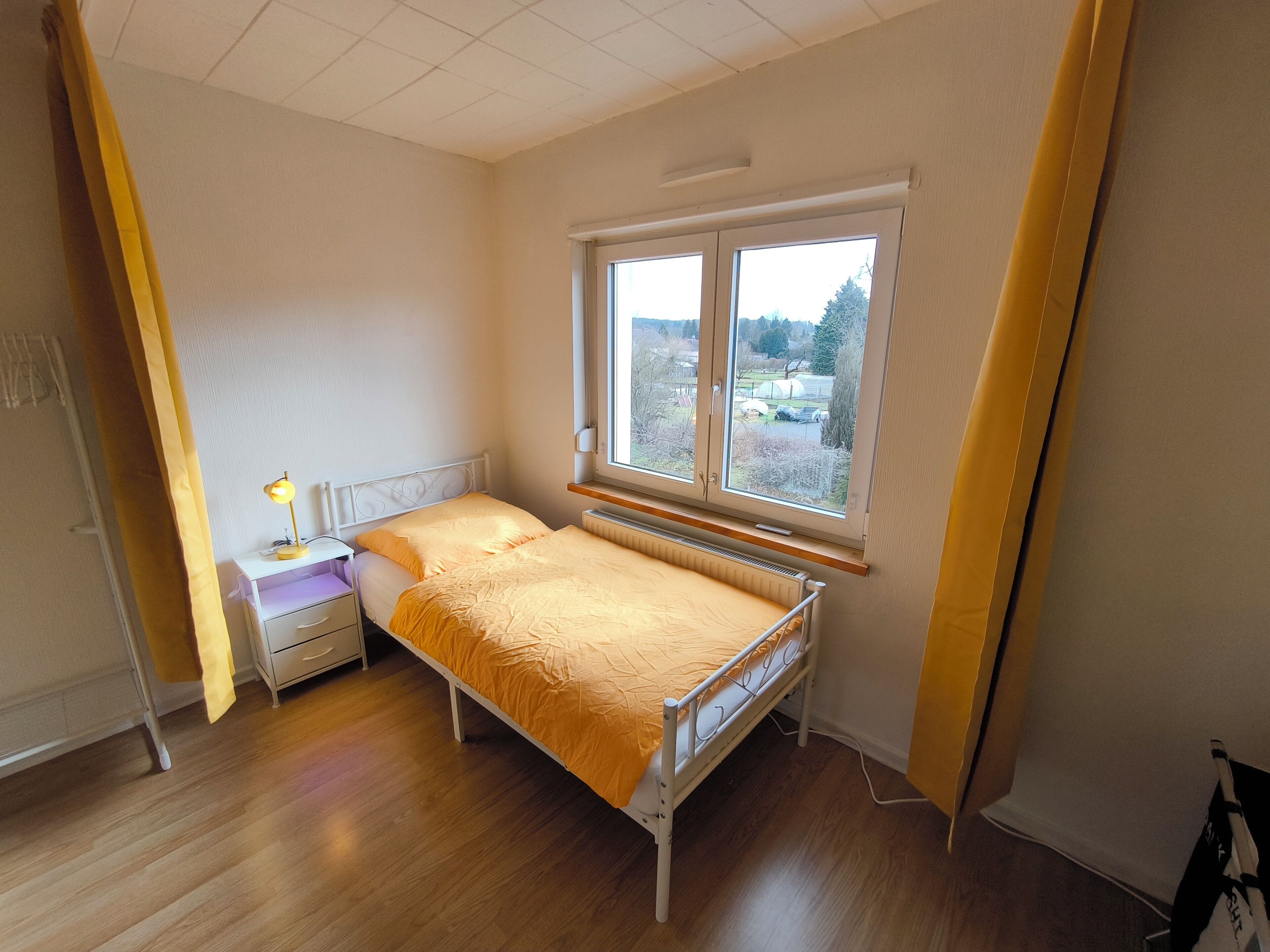 A bedroom with a bed, a nightstand, a lamp, and a window with a view of the outdoors.