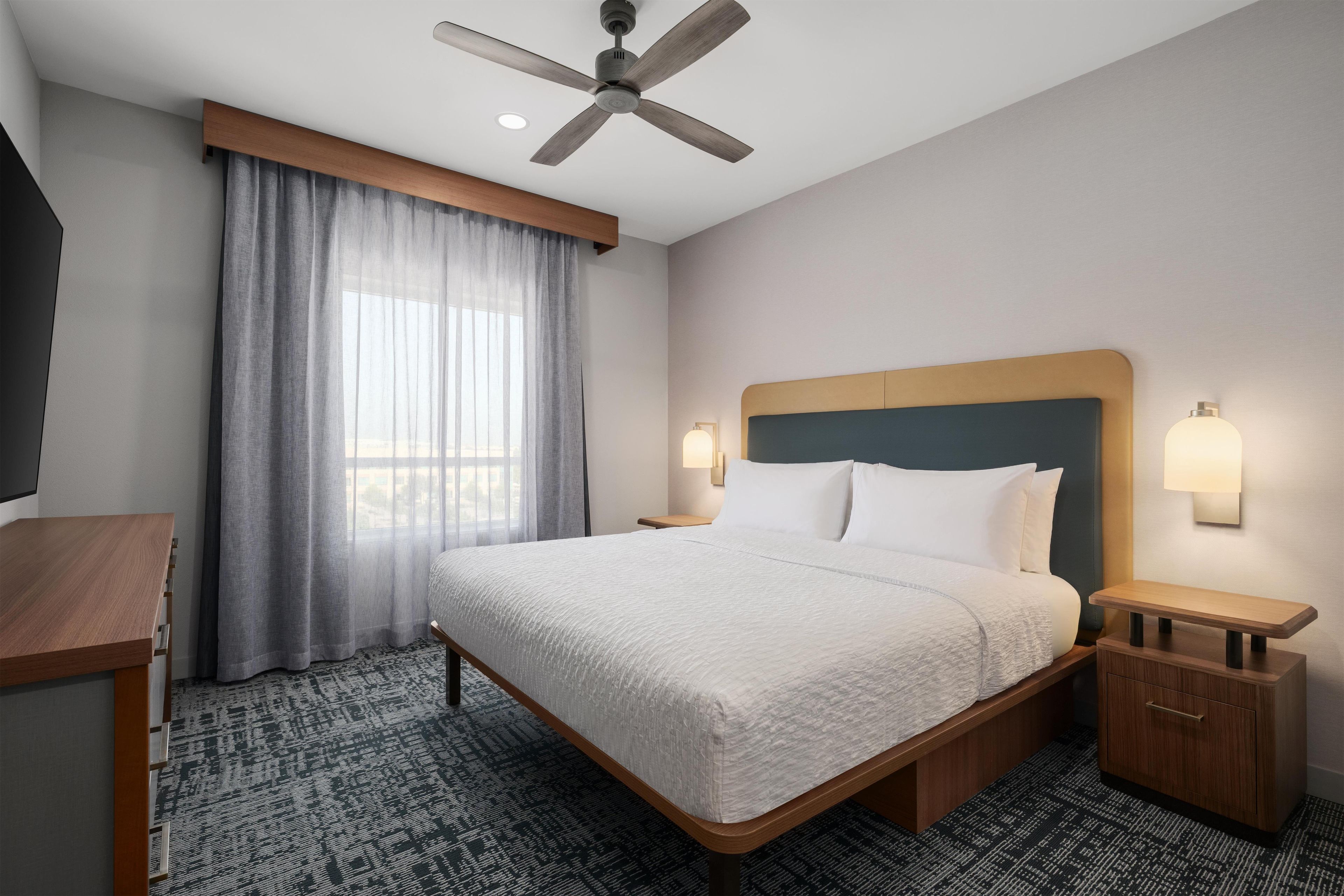 Quarto de hotel com cama, mesas de cabeceira, ventoinha no teto, janela com cortinas e luminária na parede.