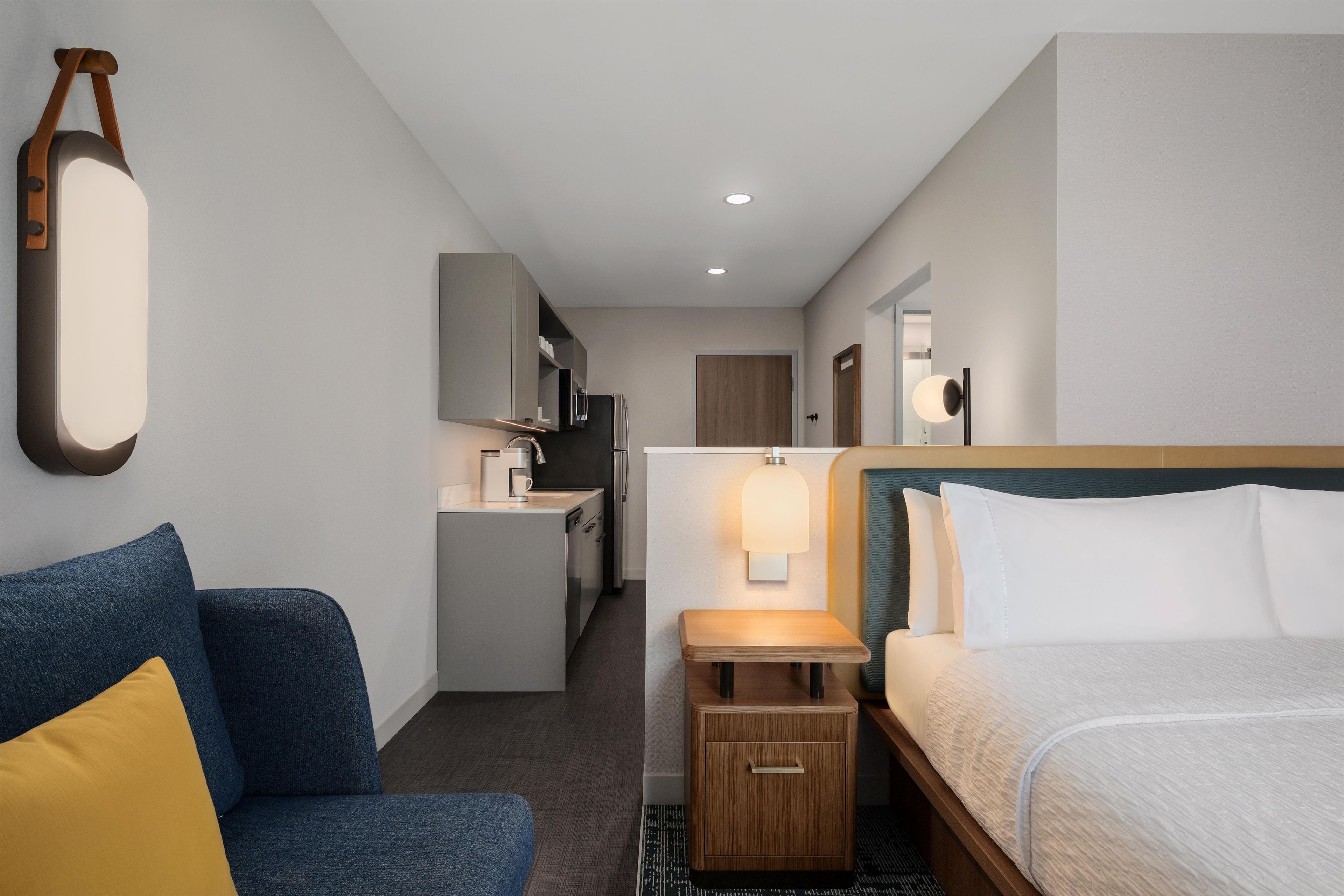 Habitación de hotel con cama, escritorio, silla, una kitchenette pequeña y un espejo.