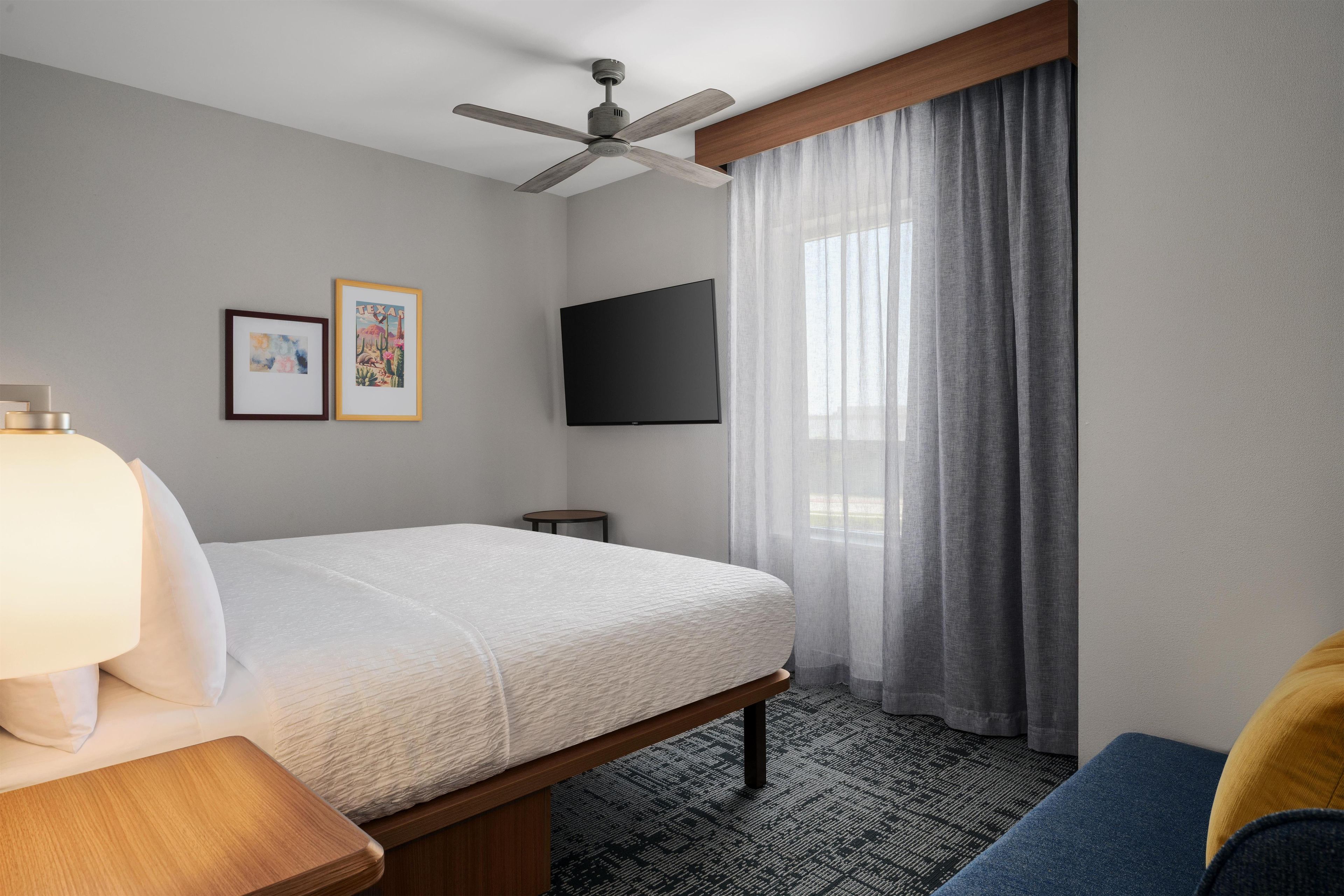A hotel room with a bed, a flat-screen TV, a ceiling fan, a window with curtains, and two framed pictures on the wall.