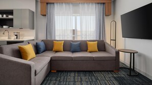 A modern hotel room with a sectional sofa, a flat-screen TV, a small round table, and a kitchen area with a coffee machine. - Homewood Suites By Hilton Coppell Dfw Airport North (Coppell)
