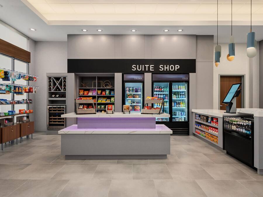 A modern hotel suite shop with a central display, refrigerated sections, and shelves stocked with various products.
