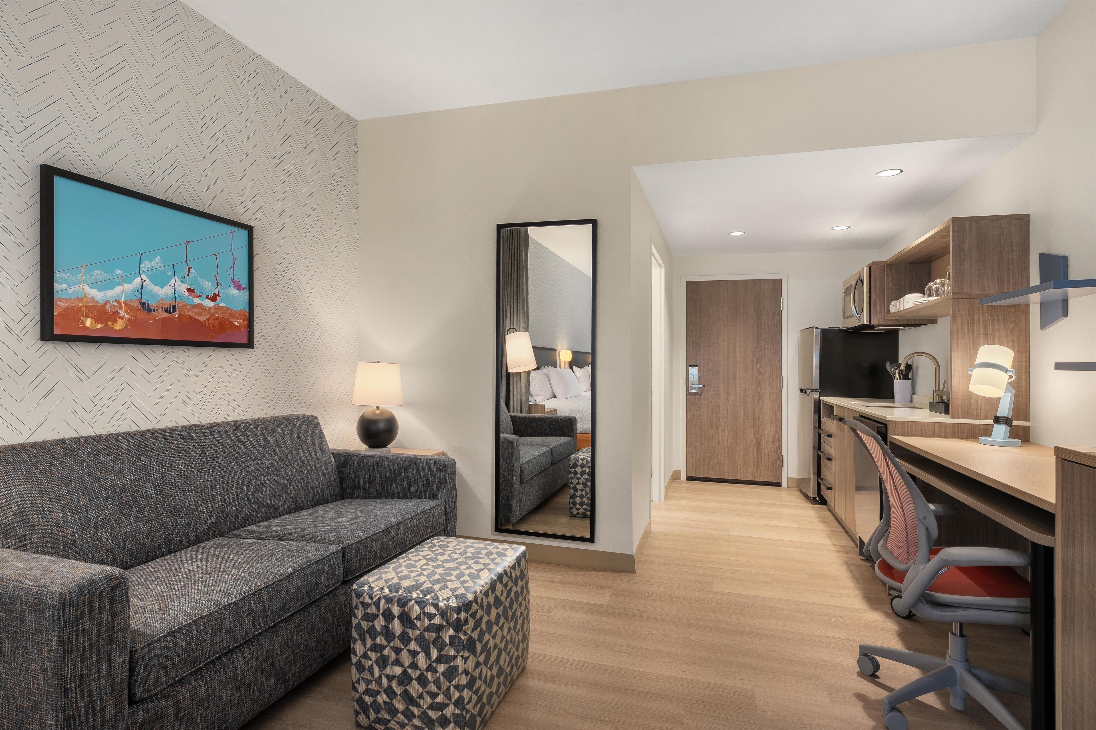 A modern hotel room with a sofa, ottoman, a painting, a mirror, a desk with a chair, and a kitchenette.