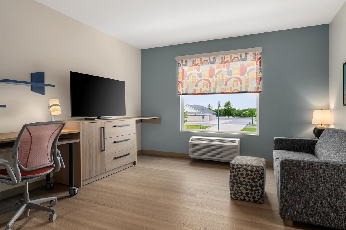 Une chambre d’hôtel moderne équipée d’un téléviseur à écran plat, d’un bureau avec une chaise, d’un canapé, d’un pouf à motifs et d’une fenêtre donnant sur un immeuble et des espaces verts.