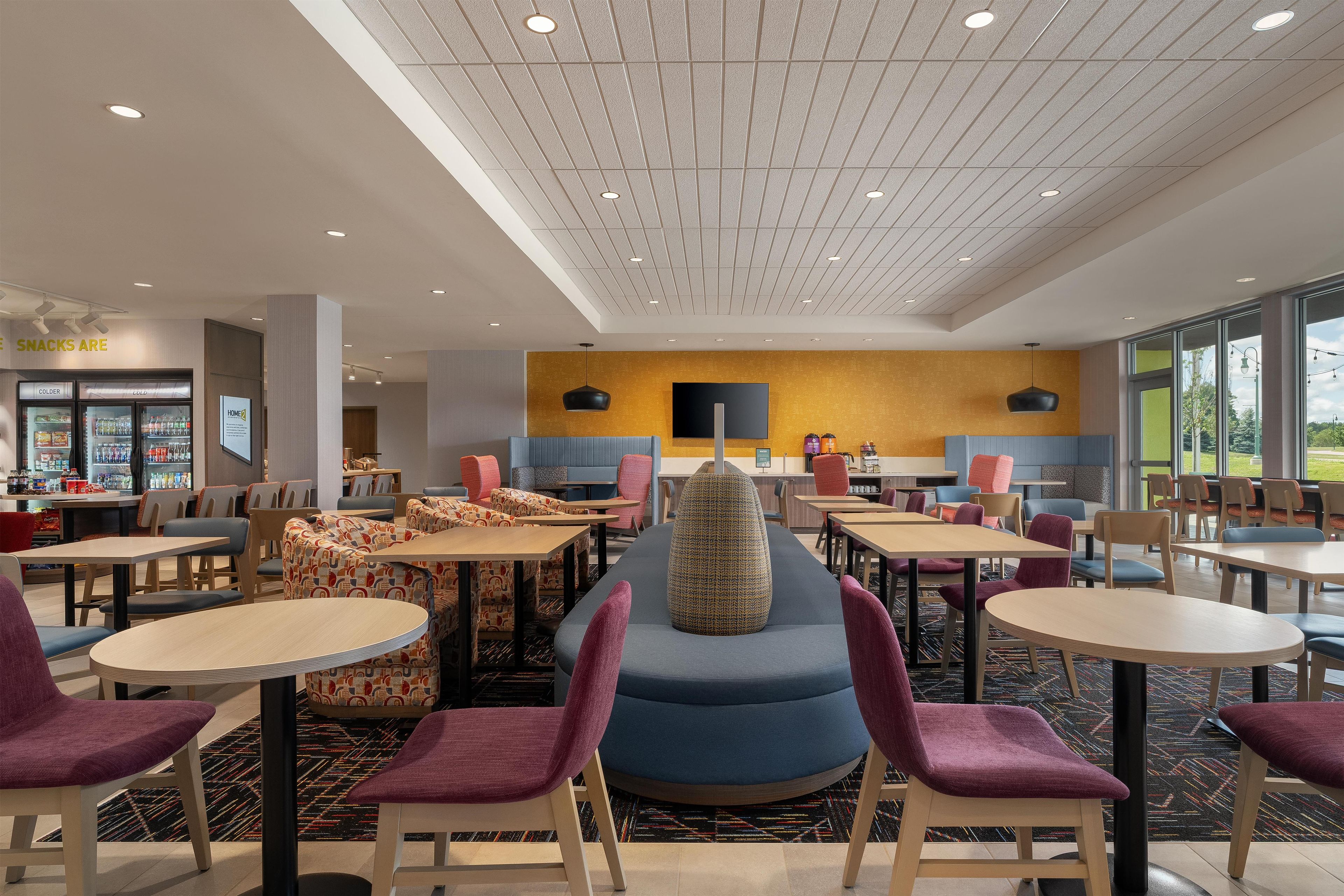 A modern dining area with multiple tables and chairs, a central seating area with a unique design, and a vending machine labeled 'SNACKS ARE'.