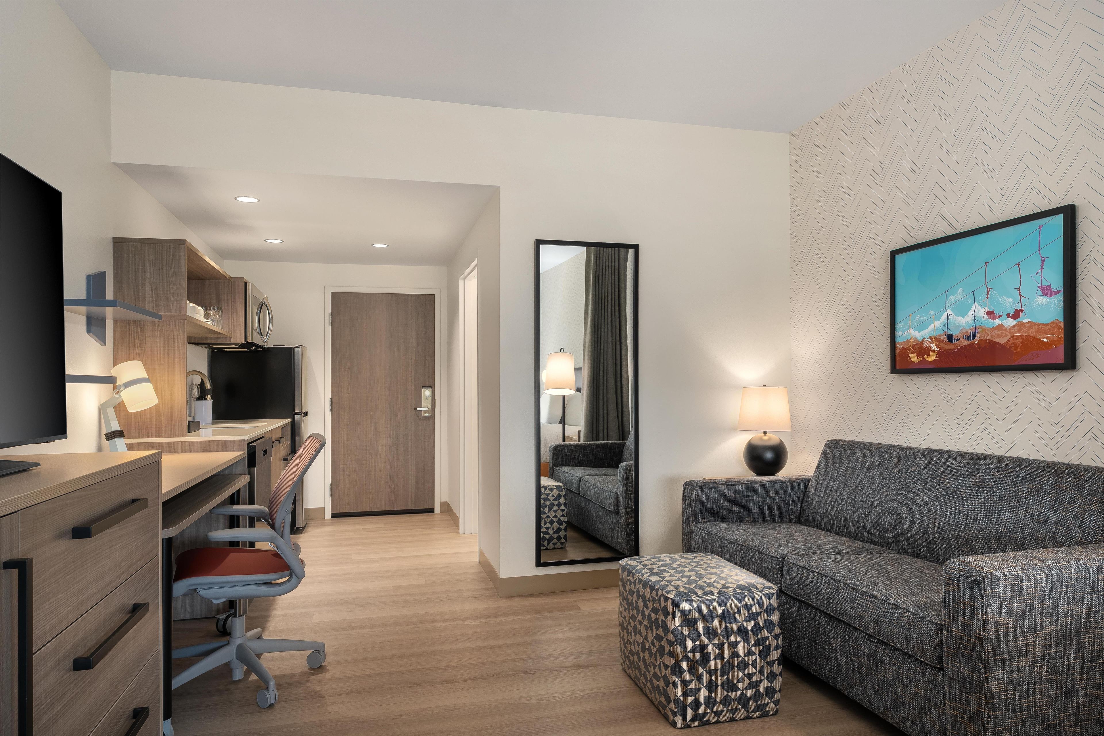 A modern hotel room with a desk, chair, sofa, ottoman, and a large mirror.