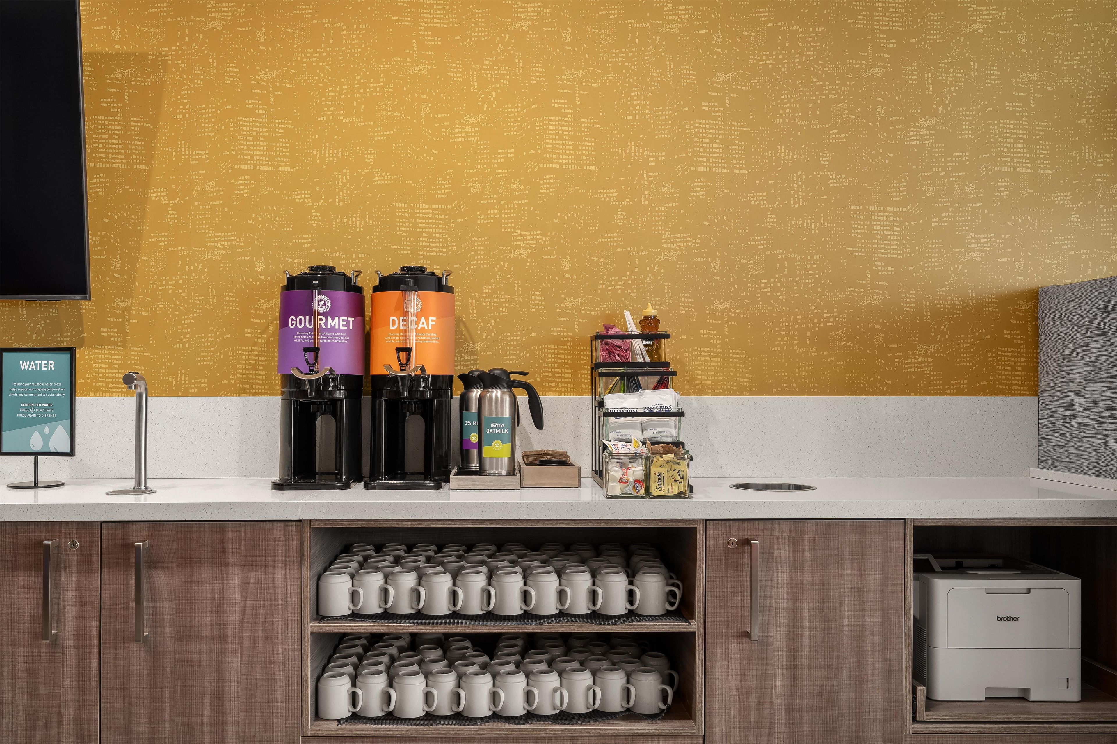 A coffee station with a water dispenser, two coffee makers, a honey dispenser, and a mug display.