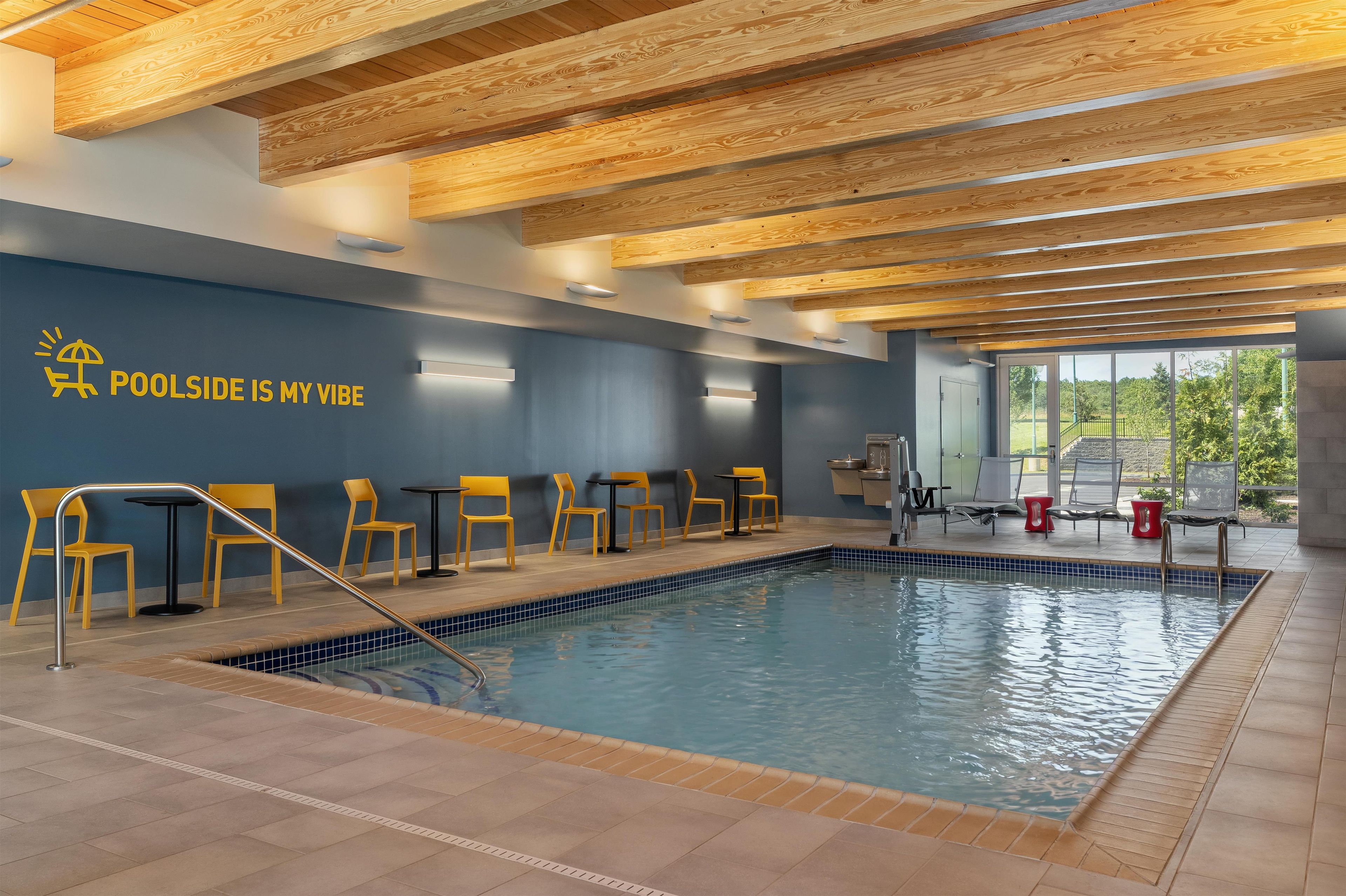 An indoor swimming pool with wooden ceiling beams, a blue wall with a sun and poolside icon, and yellow chairs.