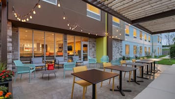 An outdoor patio with seating and tables, a stone wall, and a building with large windows in the background.