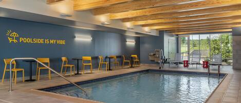 An indoor swimming pool with wooden ceiling beams, a blue wall with a sun and poolside icon, and yellow chairs.