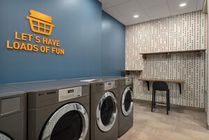 Un cuarto de lavandería con secadoras industriales y un cartel en la pared que dice 'LET'S HAVE LOADS OF FUN'.