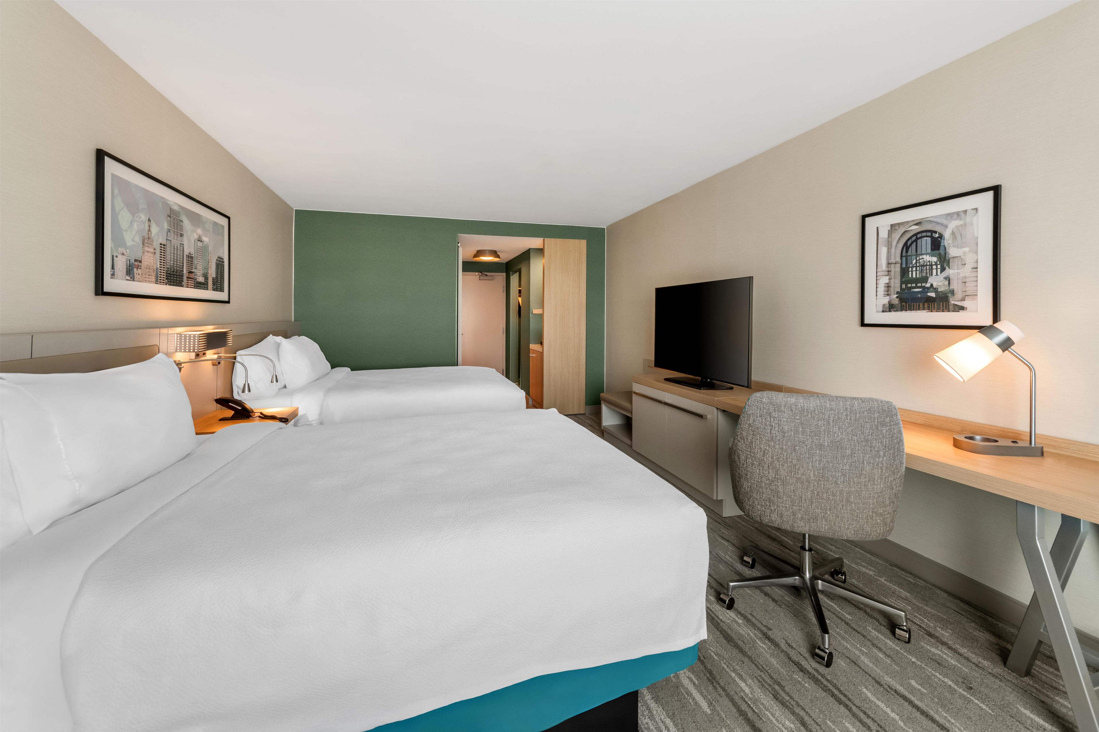 A hotel room with a large bed, a desk, a chair, a television, and two framed pictures on the walls.