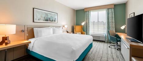 A hotel room with a large bed, a desk with a flat-screen TV, a chair, and a window with curtains.