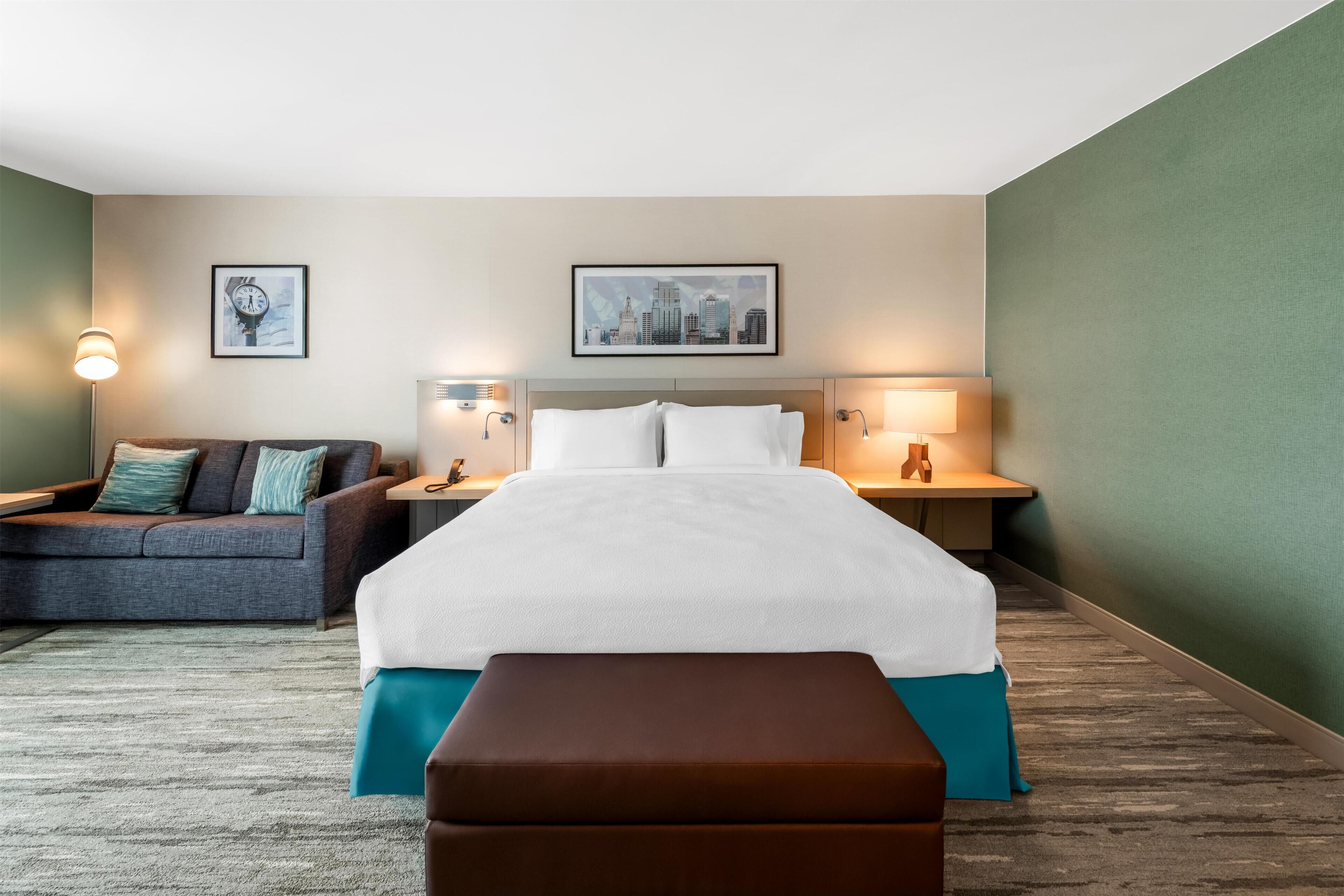 A hotel room with a bed, a sofa, two bedside tables with lamps, and two wall-mounted artworks.