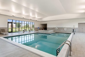 A large indoor swimming pool with clear blue water, surrounded by lounge chairs and a view of a building through large windows.