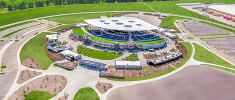 Un estadio con un campo circular central, rodeado de un área de estacionamiento y un perímetro ajardinado.