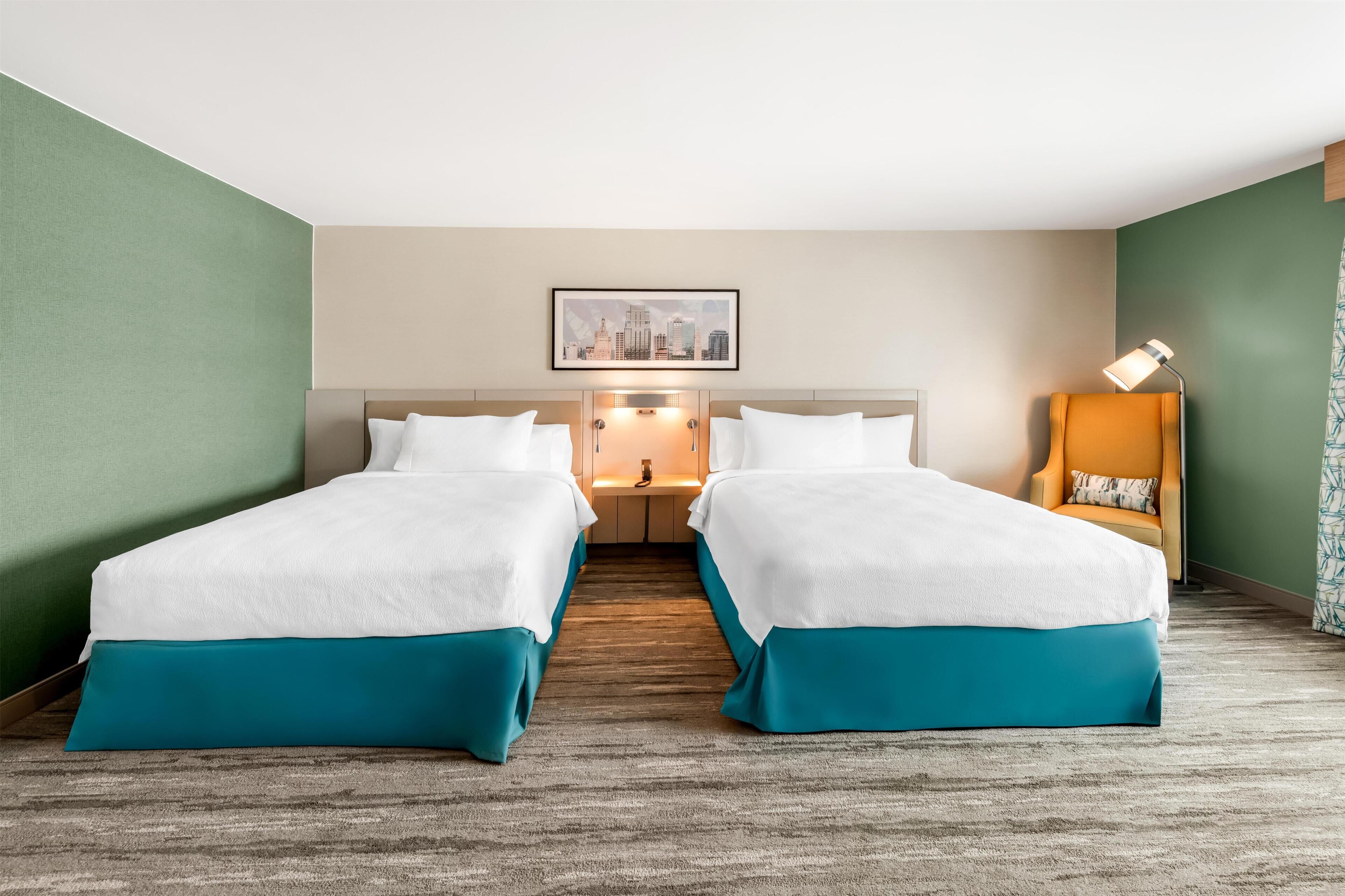 A hotel room with two beds, a nightstand, a lamp, and a framed cityscape picture on the wall.