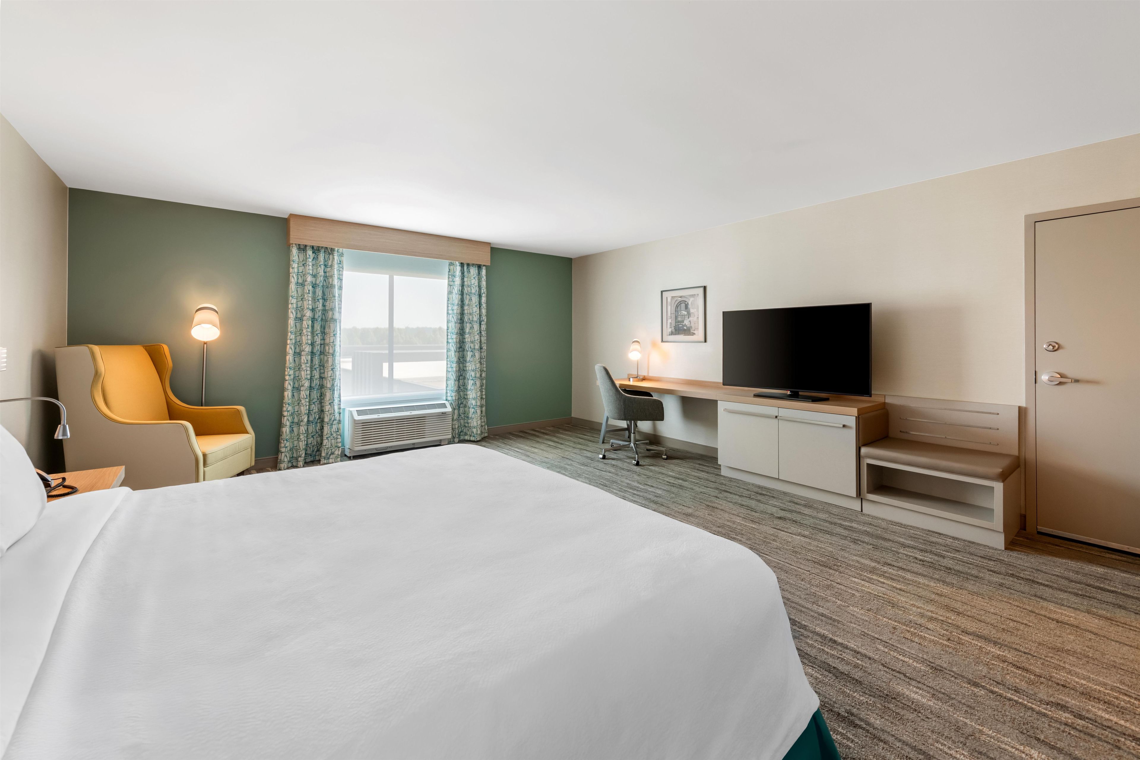 A hotel room with a large bed, a desk with a chair, a television, and a door.