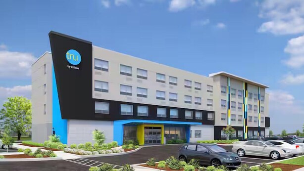 Ein modernes Hotelgebäude mit auffälligem Logo an der Seite, mehreren Etagen und einem Parkplatz mit Autos.