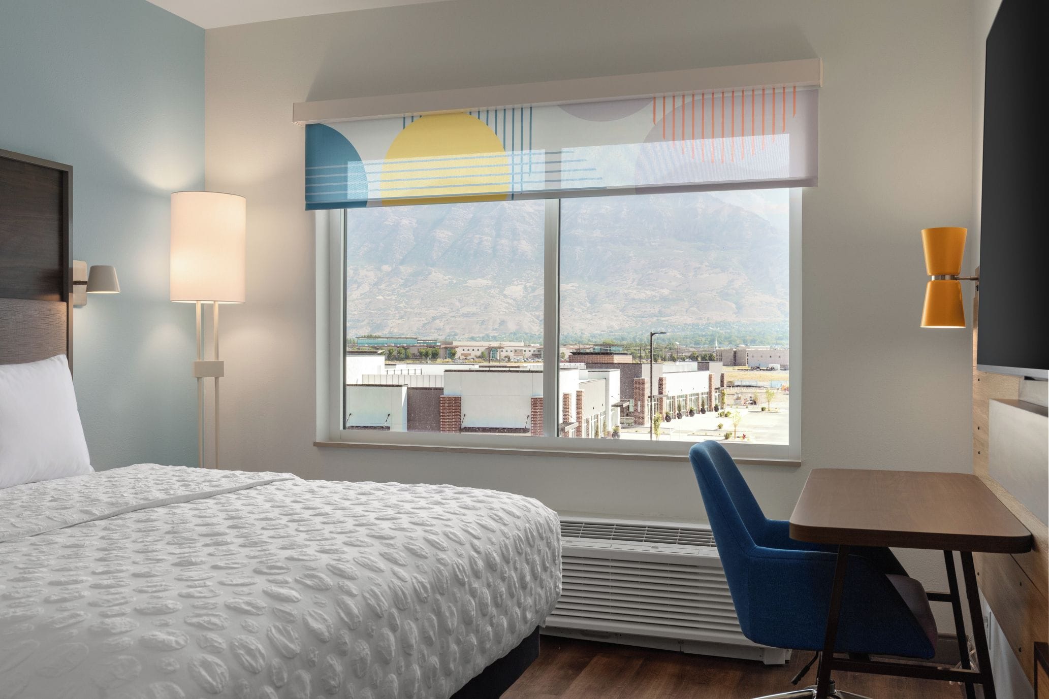 A hotel room with a large bed, a desk, a chair, a window with a view of buildings and mountains, and a wall-mounted lamp.