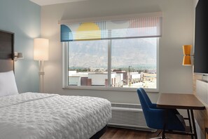 Une chambre d’hôtel avec un grand lit, un bureau, une chaise, une fenêtre donnant sur des immeubles et des montagnes, et une lampe fixée au mur.