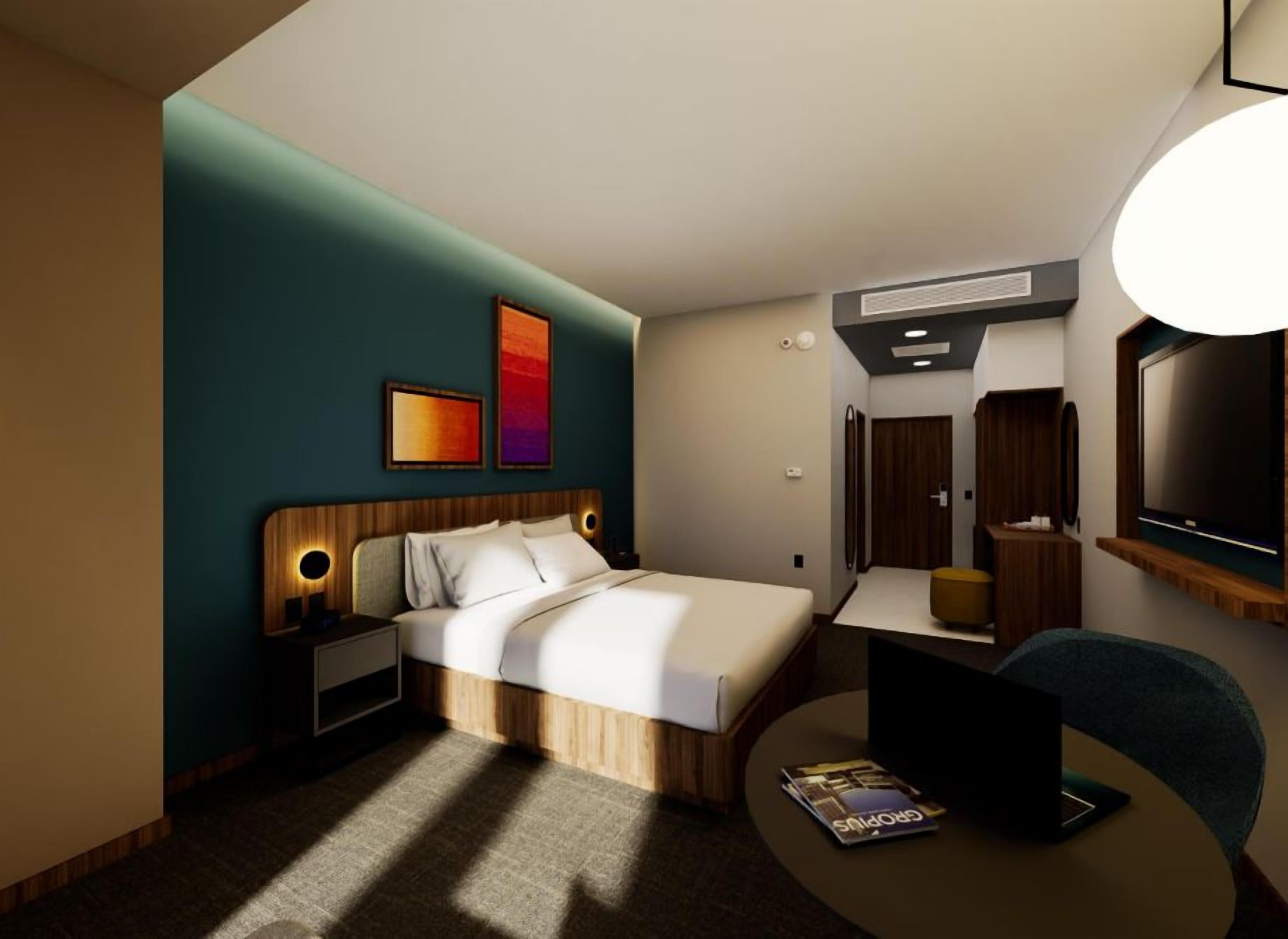 A modern hotel room with a bed, bedside table, lamp, and a desk with a chair.