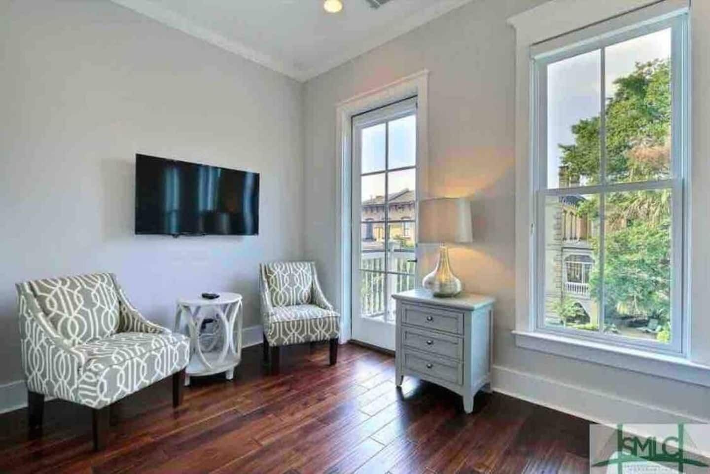 A room with hardwood flooring, two patterned armchairs, a small round table, a white side table with a lamp, a large window with a view of buildings and trees, and a flat-screen TV mounted on the wall.