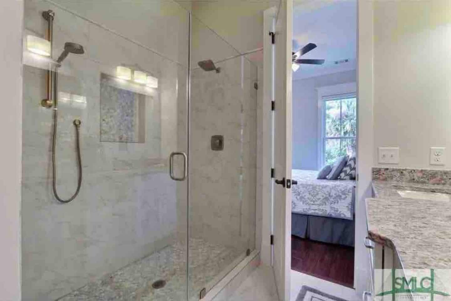 Une douche en verre avec un pommeau de douche à main et un pommeau de douche fixe, donnant sur une chambre à coucher équipée d’un lit et d’une fenêtre.