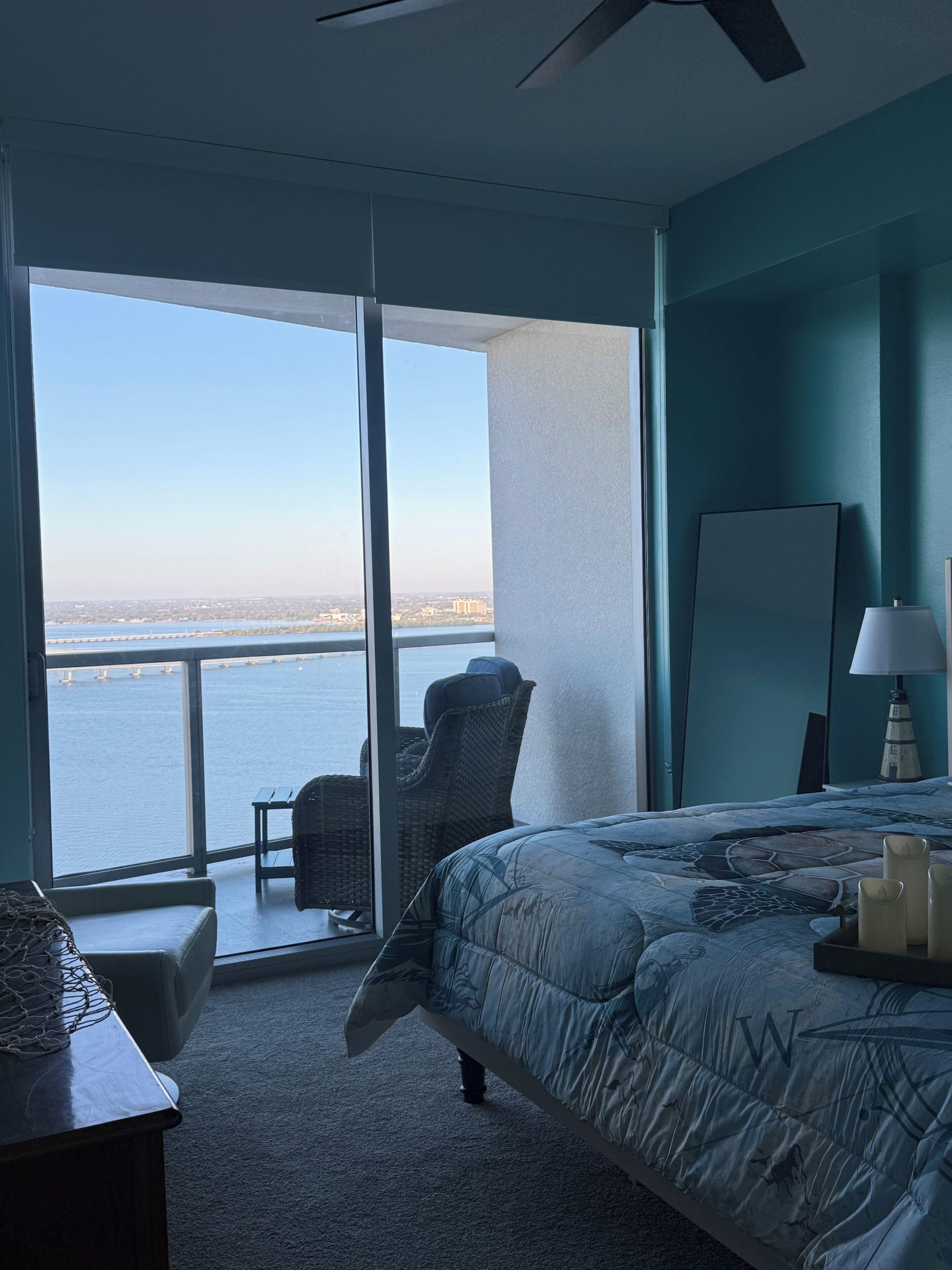 A bedroom with a large window, a bed, a chair, a desk, and a lamp.