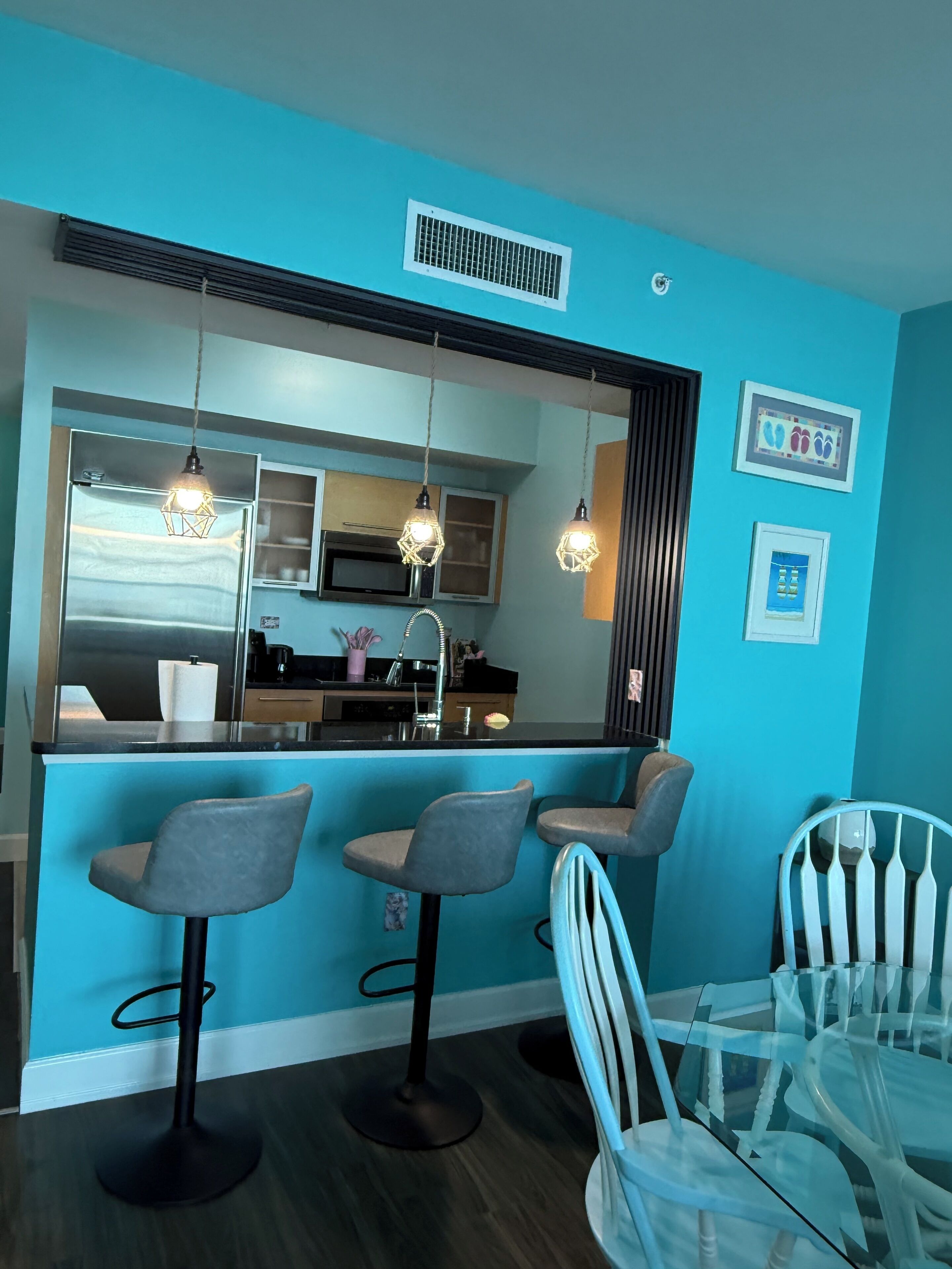 Una cocina moderna con una pared azul de acento, un refrigerador de acero inoxidable, un microondas y un lavamanos. Hay tres taburetes de bar y una mesa de comedor de vidrio con sillas.