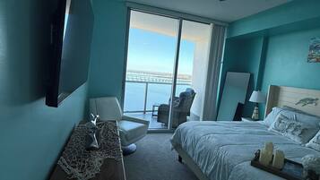 A bedroom with a bed, a desk, a chair, a ceiling fan, and a view of the water.
