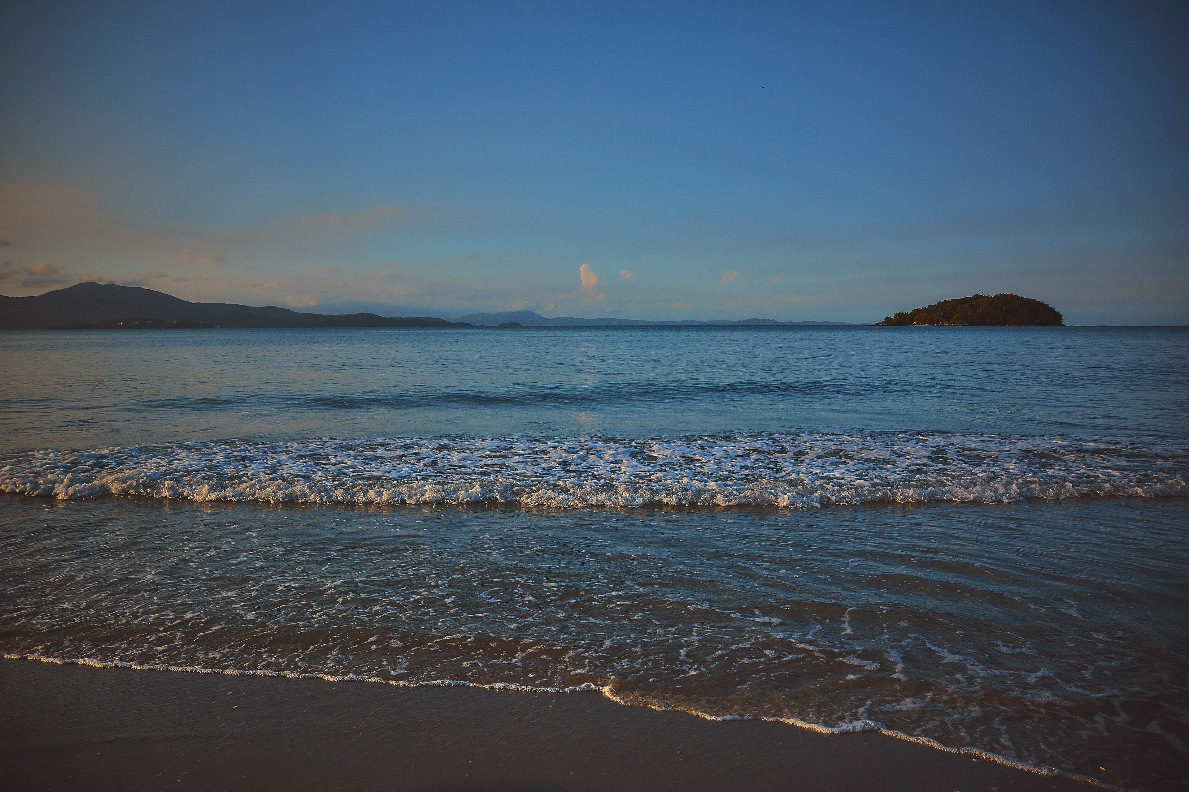 A beach with waves, a distant island, and mountains.