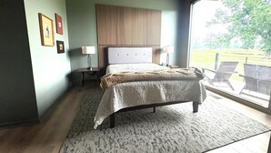 A bedroom with a large bed, a nightstand, a lamp, and a view of the outdoors.