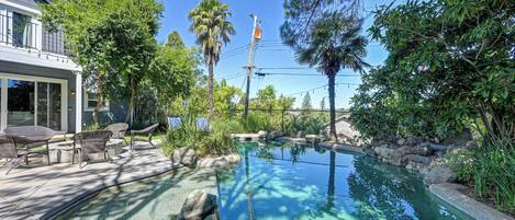 A swimming pool with clear water, surrounded by rocks and plants, and a patio area with chairs and a table.