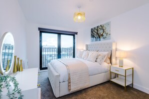 A modern bedroom with a large bed, a bedside table, a mirror, and a view of the city through a balcony. - 2BDR | Free Parking | Stylish Liverpool Apartment (Liverpool)