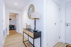 A hallway with a desk, a lamp, and a mirror. There is a door on the right and a dining area in the background. - 2BDR | Free Parking | Stylish Liverpool Apartment (Liverpool)