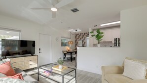 Ein modernes Wohnzimmer mit Glastisch, Sofa und Fernseher. Im Hintergrund ist ein Essbereich zu sehen, durch eine offene Tür ist die Küche erkennbar.