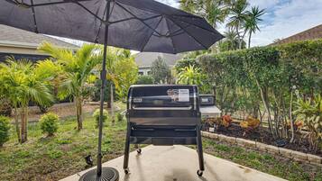 Ein Grill mit Sonnensegel, auf einer Beton-Terrasse, umgeben von Grünflächen und Palmen.