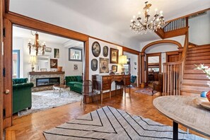 Un salón amplio con chimenea, sofá verde, escalera de madera y una araña.