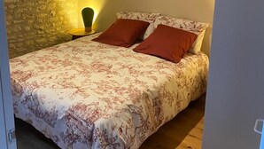 Une chambre avec un mur en pierre, un tableau représentant un paysage, un lit avec une literie à motifs floraux et une table de chevet avec une lampe.