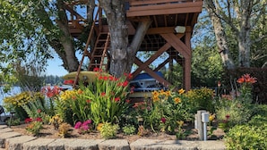Une terrasse en bois avec un bateau, entourée d’un jardin où poussent différentes sortes de fleurs et bordée d’un mur de pierres.