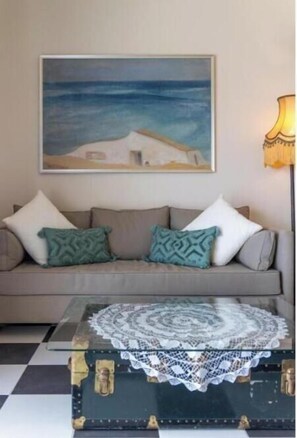 A living room with a sofa, a painting, a lamp, and a vintage suitcase with a lace cover.